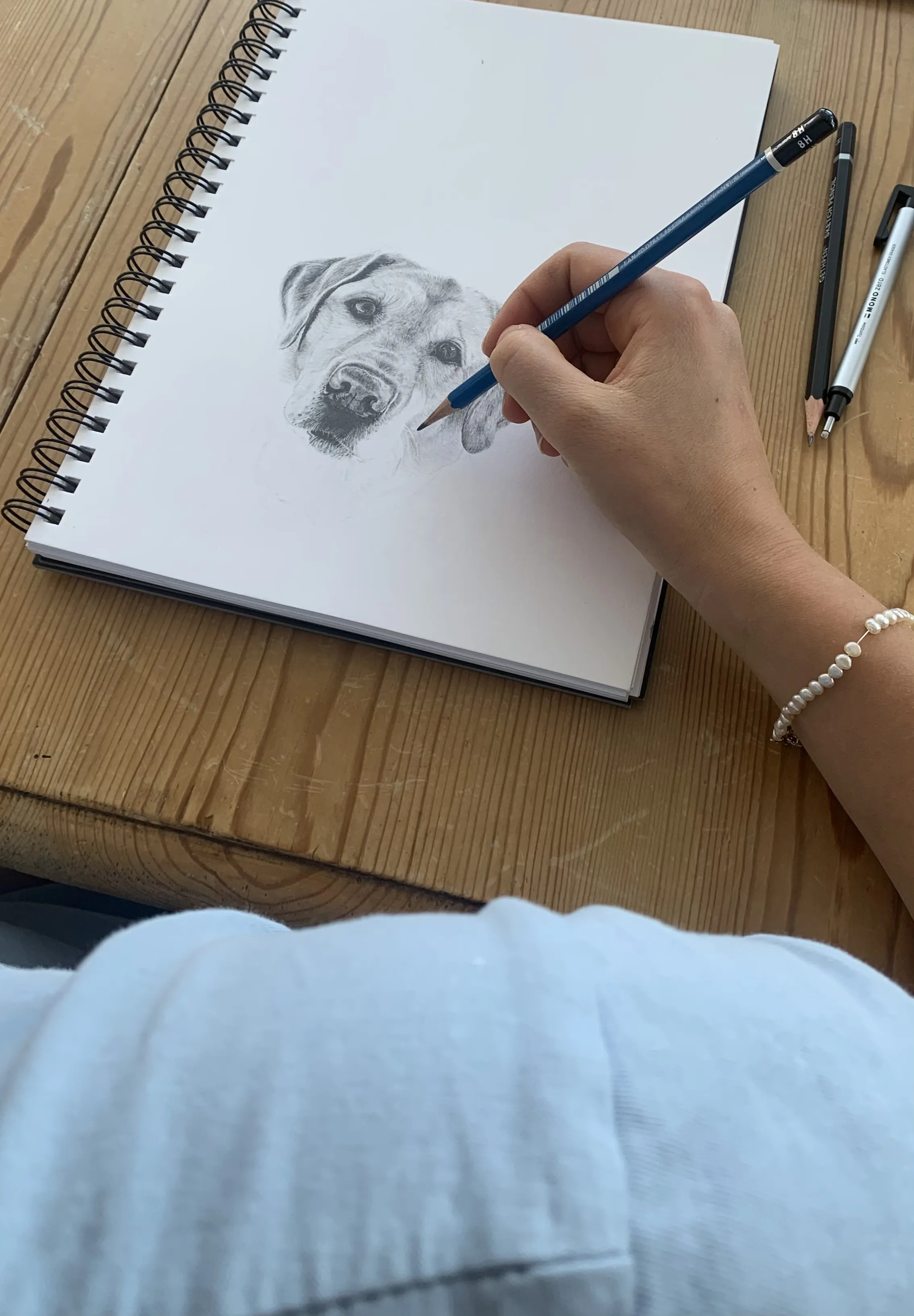 Hanna's hand at work, shading a graphite drawing of a dog in a spiral sketchbook.